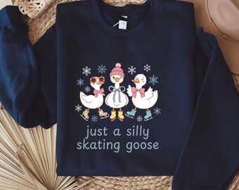 May include: Navy blue sweatshirt featuring a whimsical illustration of three white geese wearing ice skates, hats, and bows, surrounded by snowflakes. The text "just a silly skating goose" is printed below the geese.