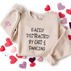 以下が含まれることがあります： 「EASILY DISTRACTED BY CATS & DANCING」の黒い文字が入ったベージュ色のスウェットシャツ。赤、ピンク、紫の色合いのハート型の装飾が周りに散りばめられています。