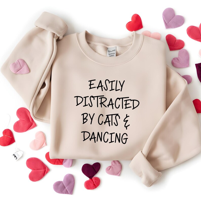 以下が含まれることがあります： 「EASILY DISTRACTED BY CATS & DANCING」の黒い文字が入ったベージュ色のスウェットシャツ。赤、ピンク、紫の色合いのハート型の装飾が周りに散りばめられています。