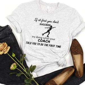 May include: Light gray t-shirt with the text "If at first you don't succeed, try doing what your coach told you to do the first time" and a silhouette of a figure skater. The shirt is paired with dark jeans and leopard print flats.