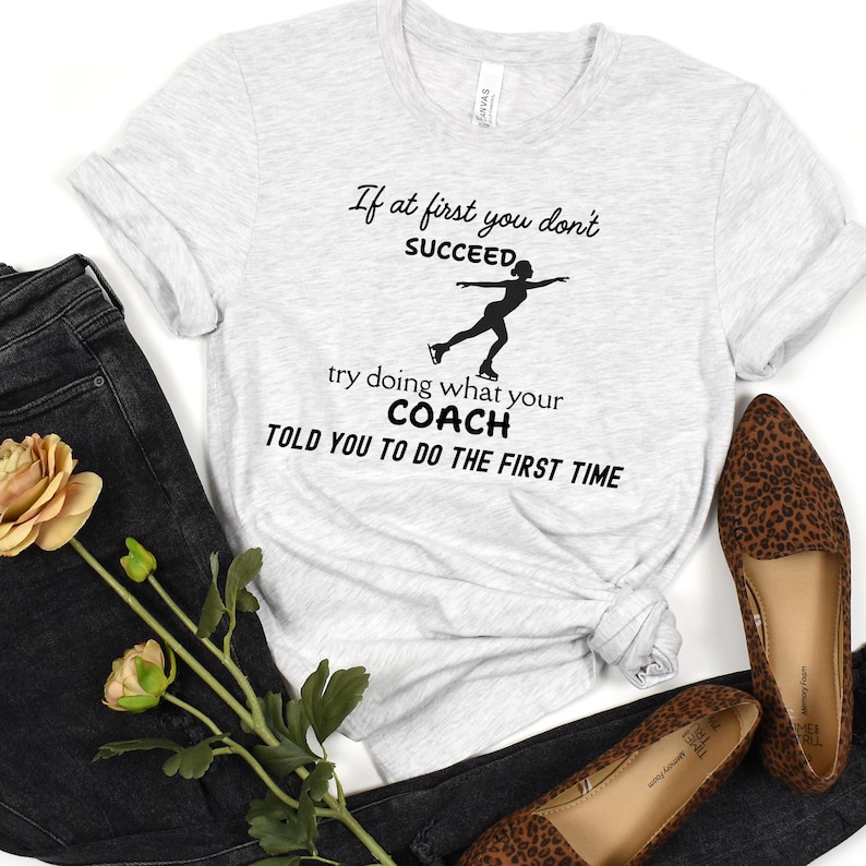 May include: Light gray t-shirt with the text "If at first you don't succeed, try doing what your coach told you to do the first time" and a silhouette of a figure skater. The shirt is paired with dark jeans and leopard print flats.