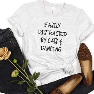 以下が含まれることがあります： 「EASILY DISTRACTED BY CATS & DANCING」の黒い文字がプリントされたライトグレーのTシャツ。黒いジーンズとレオパード柄のフラットシューズとコーディネートされています。黄色のバラと緑も画像にあります。