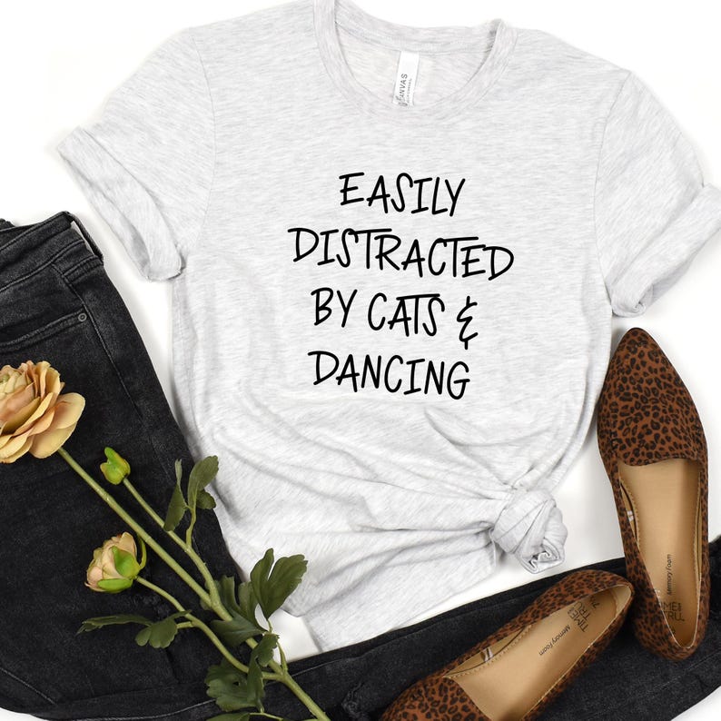 以下が含まれることがあります： 「EASILY DISTRACTED BY CATS & DANCING」の黒い文字がプリントされたライトグレーのTシャツ。黒いジーンズとレオパード柄のフラットシューズとコーディネートされています。黄色のバラと緑も画像にあります。