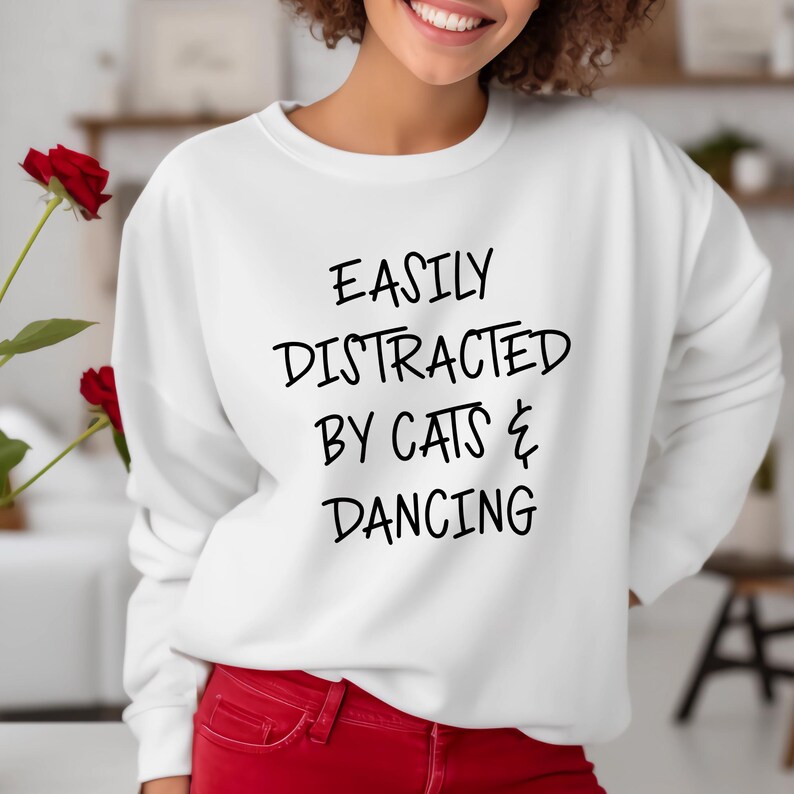 以下が含まれることがあります： 「EASILY DISTRACTED BY CATS & DANCING」の黒い文字がプリントされた白いスウェットシャツ。クルーネックのスタイルです。赤いパンツを着用しています。画像の左側には赤いバラが見えます。