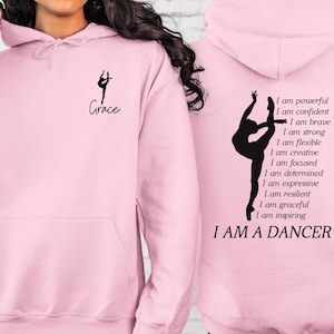 May include: Light pink hoodie with a black silhouette of a dancer on the front and back. The front features the word "Grace" and a dancer graphic. The back displays a dancer graphic and the text "I AM A DANCER" with inspirational affirmations.