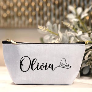 May include: A white canvas cosmetic bag with a black zip and trim. The bag features the name "Olivia" in a cursive font, with a small heart and a figure skate graphic.