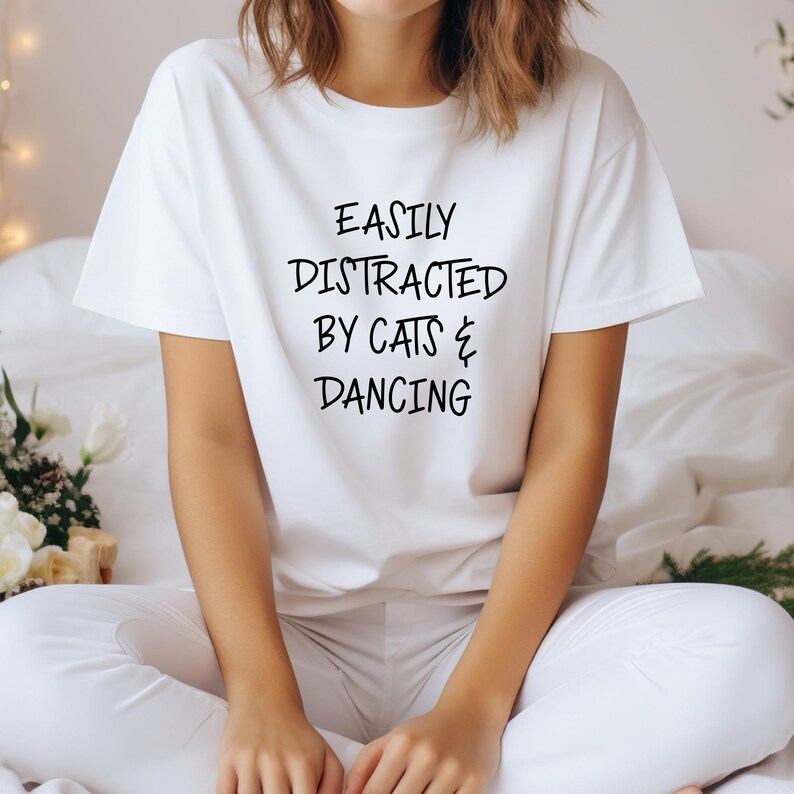 以下が含まれることがあります： 「EASILY DISTRACTED BY CATS & DANCING」の黒い文字が入った白いTシャツ。半袖で、柔らかい素材のようです。白いズボンを履いた人がベッドに座っています。