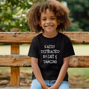 以下が含まれることがあります： 「EASILY DISTRACTED BY CATS & DANCING」の白文字がプリントされた黒いTシャツ。木製の柵の前で笑顔を見せる、巻き毛の茶色の髪の子供が着ています。