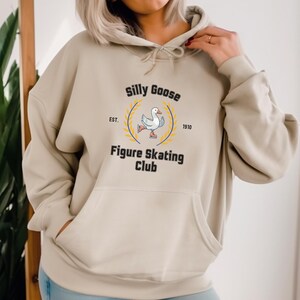 May include: A beige hoodie with a graphic of a goose ice skating. The graphic says "Silly Goose Figure Skating Club" with "Est. 1910" underneath.