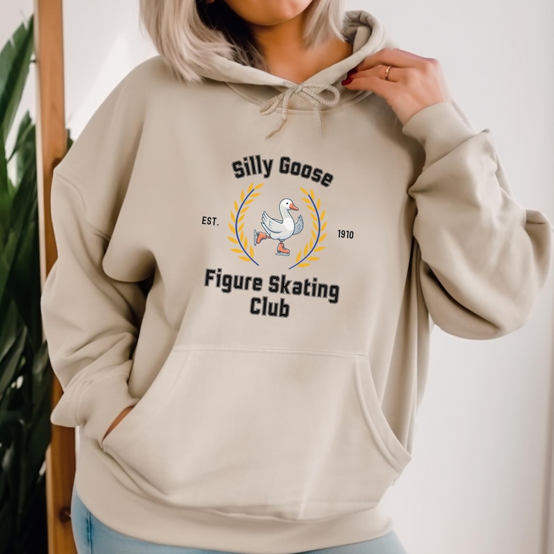 May include: A beige hoodie with a graphic of a goose ice skating. The graphic says "Silly Goose Figure Skating Club" with "Est. 1910" underneath.