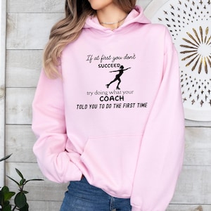 May include: A light pink hoodie with a black graphic of a figure skater and text that reads "If at first you don't SUCCEED, try doing what your COACH TOLD YOU TO DO THE FIRST TIME."