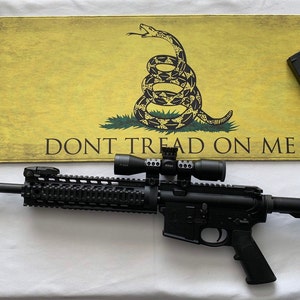 May include: A black rifle with a scope and a magazine on a yellow and green "Don't Tread On Me" flag background.
