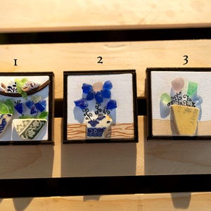 May include: Three framed mosaic art pieces featuring floral arrangements in decorative vases. The mosaics use blue, green, and yellow glass pieces. Each piece is set against a white background and framed in a dark wood.