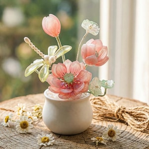 May include: A miniature white ceramic vase displays an artificial flower arrangement. The flowers are predominantly pink and peach, with green leaves and stems. The arrangement is on a rustic wooden surface, with small white daisies scattered around.