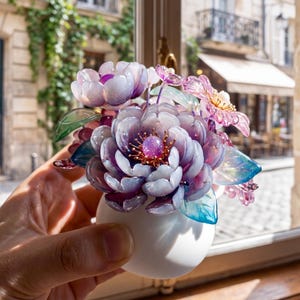 以下が含まれることがあります： 小さな白い花瓶に、造花のブーケが入っています。花は紫、ピンク、青の様々な色合いで、緑の葉が付いています。花瓶は窓の前に置かれ、背景には街並みが見えます。