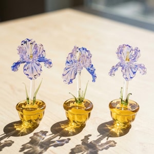 Puede incluir: Tres flores de iris de cristal en macetas color ámbar. Las flores tienen pétalos morados y azules y tallos blancos. Las macetas son redondas con una pequeña abertura. La imagen está tomada con luz natural, proyectando sombras.