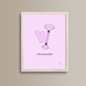 May include: A framed print with a pink background featuring a pink facial roller and a pink gua sha tool. The text "roll out your face" is written in black below the tools.