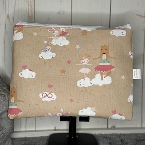 Könnte beinhalten: Eine beige Reißverschlusstasche mit einem verspielten Design mit Ballerinas, Hasen und Wolken. Die Tasche hat ein rosa, weißes und braunes Farbschema mit goldenen Sternen, die über die gesamte Fläche verteilt sind. Der Text "Handmade by Wundermaus" ist auf einem weißen Etikett gedruckt, das an der Tasche befestigt ist.