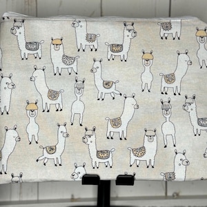 Könnte beinhalten: Eine weiße Reißverschlusstasche mit einem sich wiederholenden Muster von Lamas in verschiedenen Posen. Die Lamas sind schwarz umrandet und haben braune Akzente.