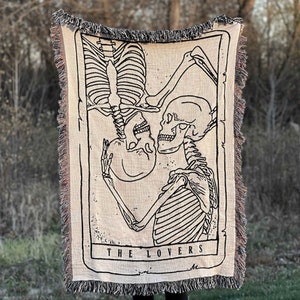 May include: A woven tapestry throw blanket featuring a black and white illustration of two skeletons embracing. The skeletons are in a tarot card style frame with the text "THE LOVERS" at the bottom.