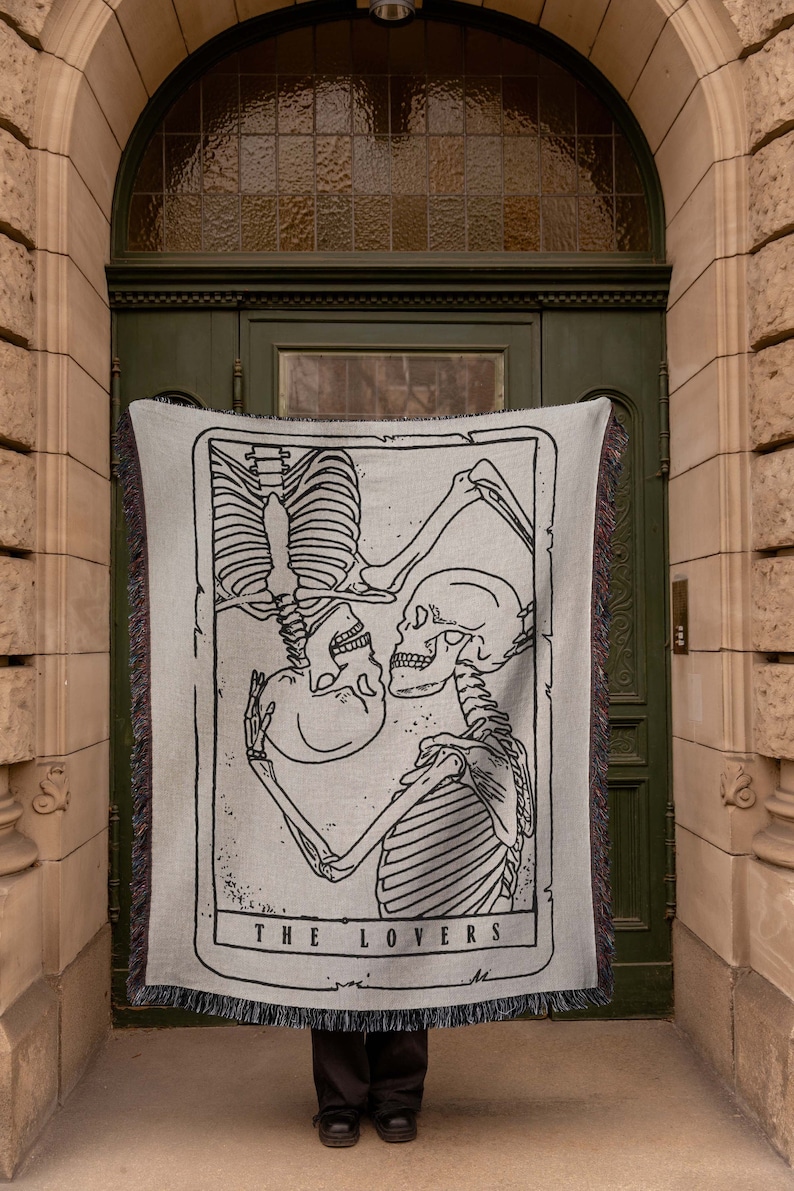 May include: A woven tapestry wall hanging featuring a black and white illustration of two skeletons embracing. The tapestry has a light gray background and the text "THE LOVERS" is printed at the bottom.