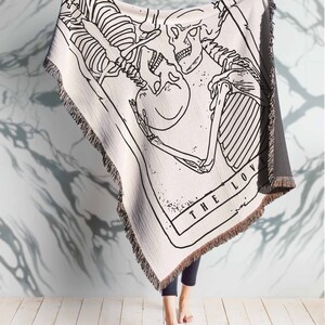 May include: A white woven tapestry throw blanket with a black and white illustration of two skeletons embracing. The text "THE LOV" is visible at the bottom of the illustration.
