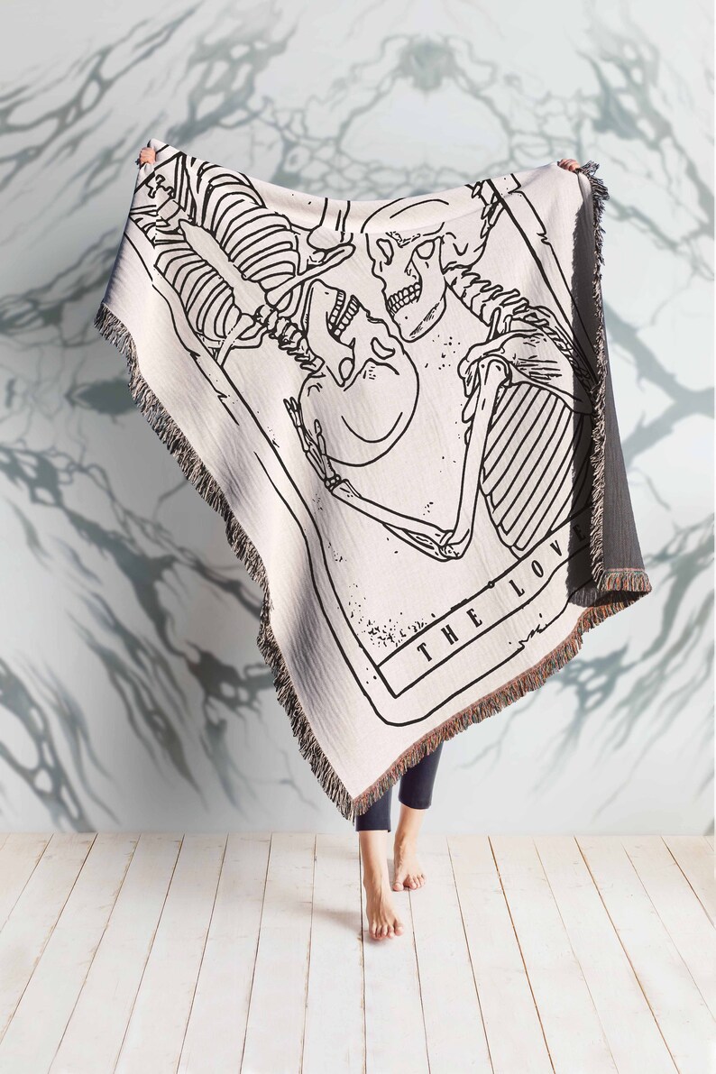 May include: A white woven tapestry throw blanket with a black and white illustration of two skeletons embracing. The text "THE LOV" is visible at the bottom of the illustration.