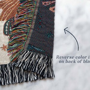 May include: A woven tapestry throw blanket with a colorful fringe. The front of the blanket features a white background with a design in brown, orange, and yellow. The back of the blanket features a reverse color image with a dark blue background.