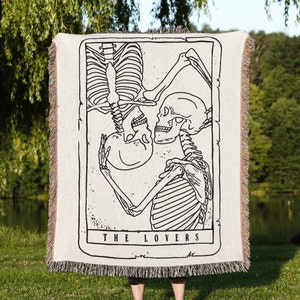 May include: A woven tapestry throw blanket featuring a black and white illustration of two skeletons embracing. The text "THE LOVERS" is printed below the image.