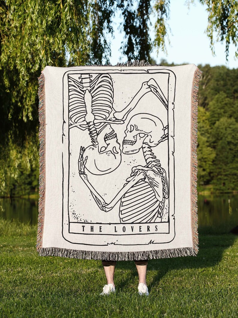 May include: A woven tapestry throw blanket featuring a black and white illustration of two skeletons embracing. The text "THE LOVERS" is printed below the image.