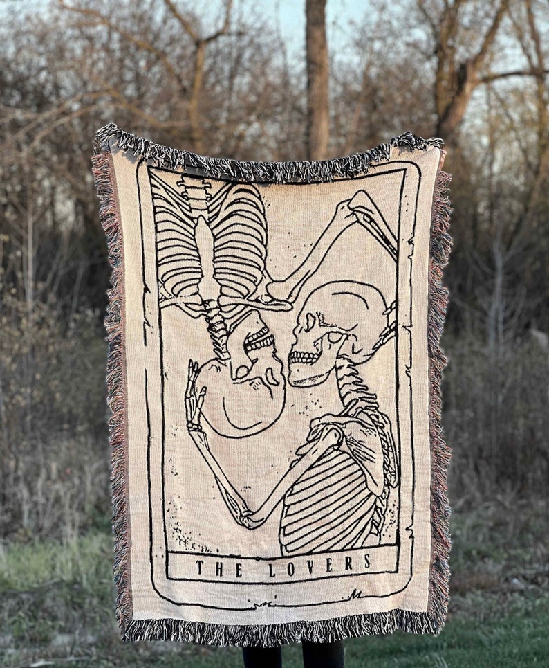 May include: A woven tapestry throw blanket featuring a black and white illustration of two skeletons embracing. The skeletons are in a tarot card style frame with the text "THE LOVERS" at the bottom.