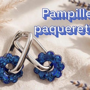 May include: A pair of silver-toned hoop earrings featuring detachable flower-shaped charms. The charms are deep blue with a glittery, textured finish. The text "Pampille paquerette" is displayed in blue letters in the upper right corner.