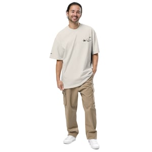 Könnte beinhalten: Eine Person trägt ein hellbeiges T-Shirt mit einem schwarzen Grafikdesign und eine beige Cargohose.