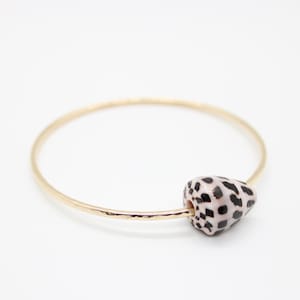 May include: A gold-colored bangle bracelet with a heart-shaped bead. The bead is white with black spots. The bracelet is on a white background.