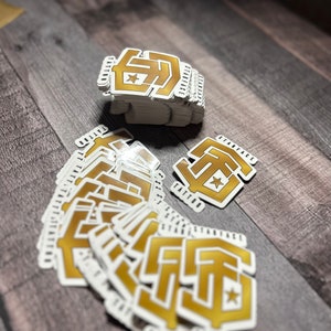 Puede incluir: Una pila de pegatinas doradas y blancas con un logotipo que presenta una estrella y el texto "STARFACE" y "TRUE GRIT" en una fuente estilizada.