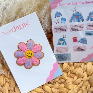 May include: A pink and purple daisy-shaped iron-on patch with a yellow smiley face center and gold trim. The patch is on a white card with the brand name "Noa Jayne". Instructions for ironing the patch are also visible.
