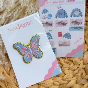 May include: A blue and pink butterfly iron-on patch with gold trim, packaged with instructions. The patch is on a white card with the brand name "Noa Jayne". The instructions show how to apply the patch to clothing.