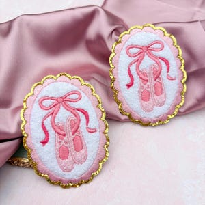 May include: Two oval-shaped patches with embroidered ballet slippers. The patches have a white center with pink ballet shoes and a pink bow. The oval is framed with a gold scalloped edge and a pink border. The background is a pink satin fabric.