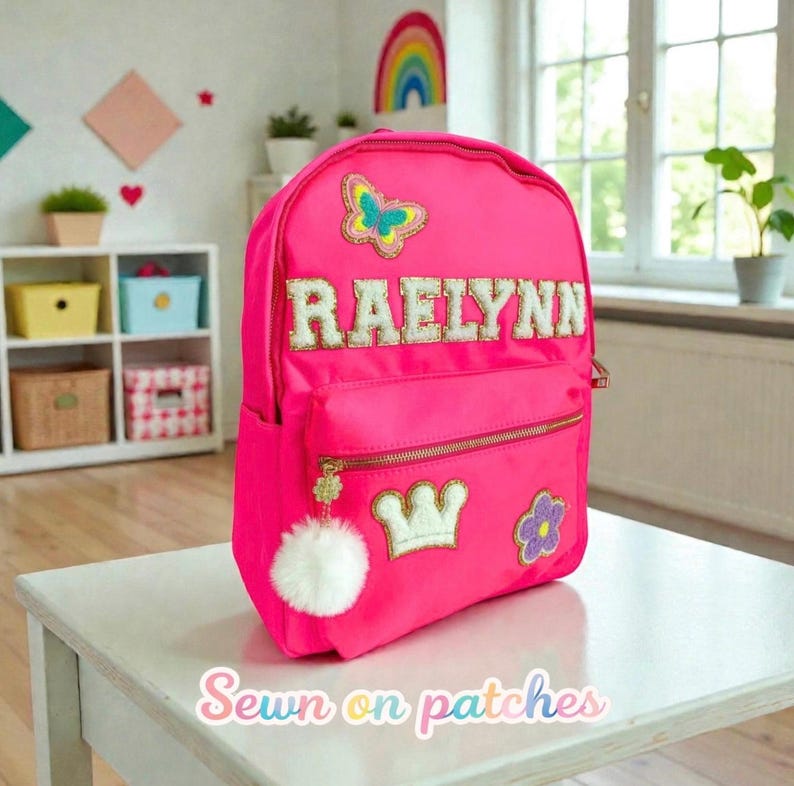 Op de afbeelding: Felroze rugzak met de naam "RAELYNN" in gouden letters. De rugzak heeft een vlinder, kroon en bloem patch. Een witte pompom-bedel hangt aan de rits. De tekst "Sewn on patches" staat onderaan.