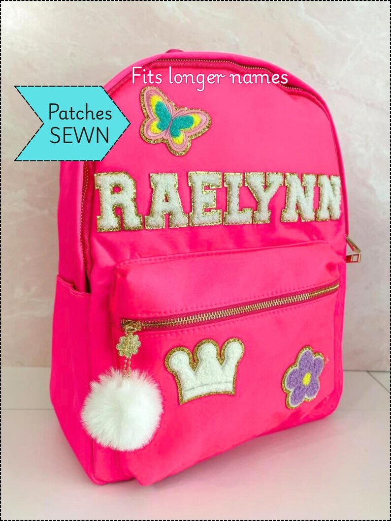 Op de afbeelding: Felroze rugzak met gouden ritsen en een witte pompon-bedel. De rugzak heeft de naam "RAELYNN" in wit, geborduurde patches van een vlinder, kroon en bloem. Tekst op de afbeelding luidt "Patches SEWN" en "Fits longer names."