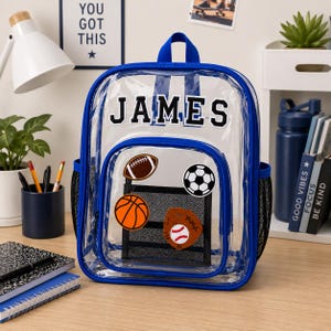 May include: A clear plastic backpack with blue trim and a top handle. The name JAMES is printed in black block letters at the top. The front pocket features a black mesh panel decorated with patches of a football, soccer ball, basketball, and glove.