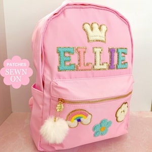 Op de afbeelding: Roze rugzak met gouden ritsen en een pluizige witte pompon-bedel. De rugzak heeft de naam "ELLIE" in kleurrijke, getextureerde letters, een kroonpatch en geborduurde regenboog- en bloempatches.