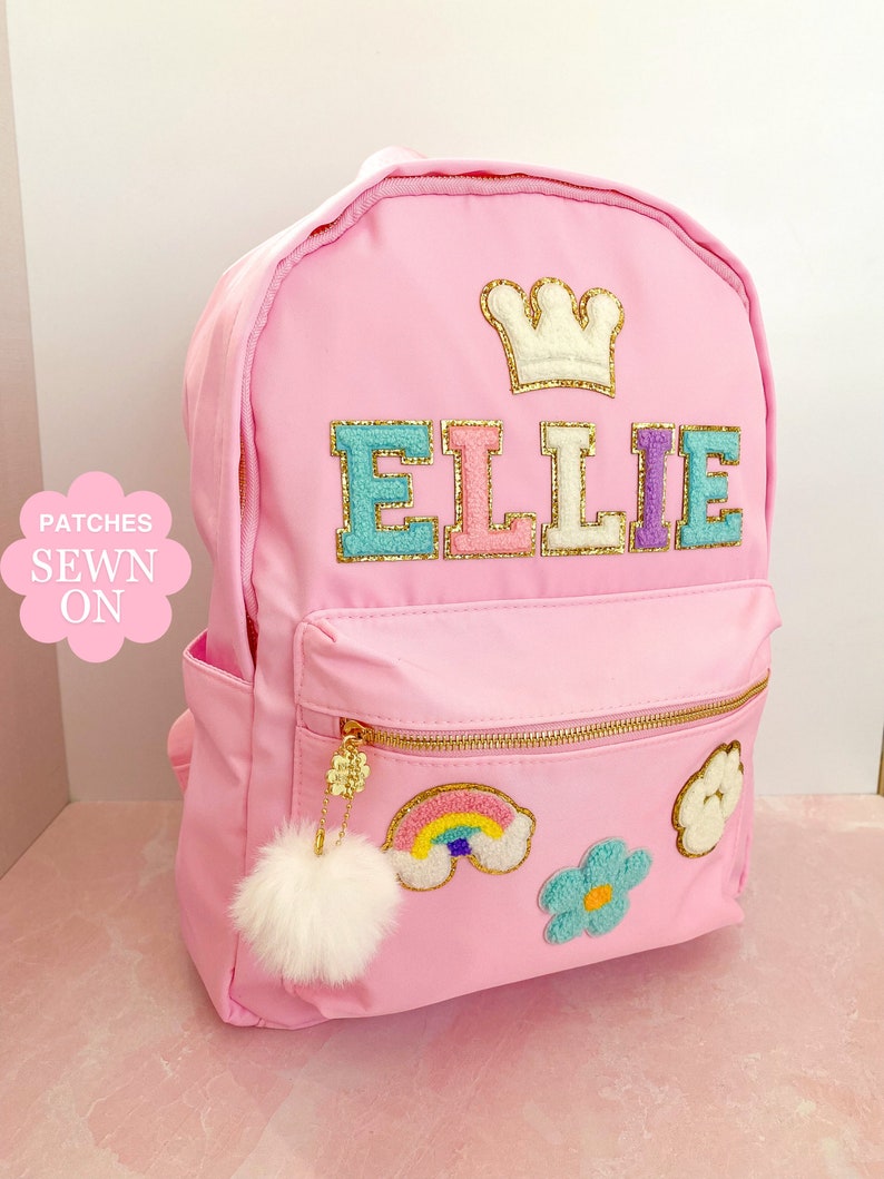 Op de afbeelding: Roze rugzak met gouden ritsen en een pluizige witte pompon-bedel. De rugzak heeft de naam "ELLIE" in kleurrijke, getextureerde letters, een kroonpatch en geborduurde regenboog- en bloempatches.