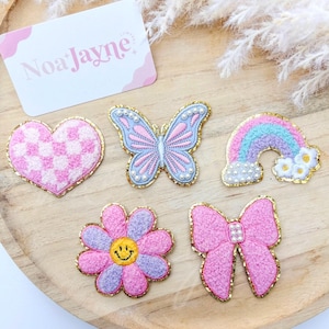 Może przedstawiać: Kolekcja haftowanych naszywek w różnych kształtach: różowo-białe serce w kratkę, niebiesko-różowy motyl, tęcza, różowy kwiat z uśmiechniętą buzią i różowa kokarda. Każda naszywka ma złote obramowanie i jest prezentowana na drewnianej powierzchni. Obecna jest również wizytówka z napisem "Noa Jayne".