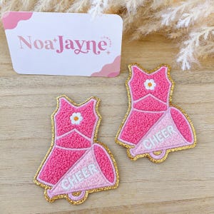 May include: Two embroidered patches shaped like cheerleading uniforms with a megaphone. Each patch features a pink textured bodice with a white flower, a pink skirt, and a light pink megaphone with the word CHEER in white letters, all outlined in gold.