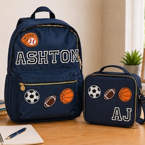 May include: A navy blue backpack and matching lunch bag set featuring gold zippers and sports-themed patches including a baseball glove, soccer ball, football, and basketball. The backpack is personalized with the name ASHTON and the lunch bag with AJ.