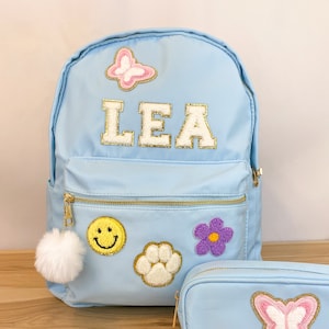Op de afbeelding: Lichtblauwe rugzak en etui set. De rugzak heeft de naam "LEA" in goudkleurige letters, plus patches van een vlinder, smiley, pootafdruk en bloem. Een witte pompon hangt aan de rits. De etui heeft een vlinder patch.