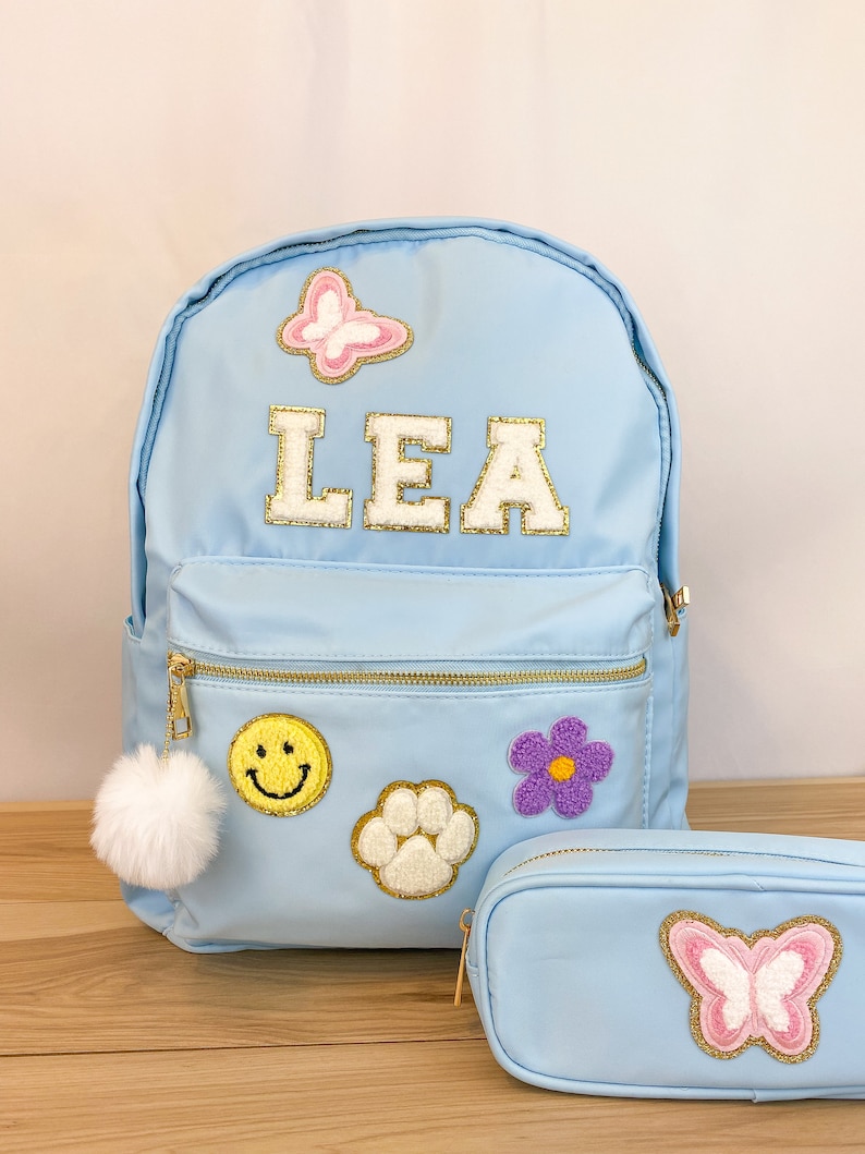 Op de afbeelding: Lichtblauwe rugzak en etui set. De rugzak heeft de naam "LEA" in goudkleurige letters, plus patches van een vlinder, smiley, pootafdruk en bloem. Een witte pompon hangt aan de rits. De etui heeft een vlinder patch.