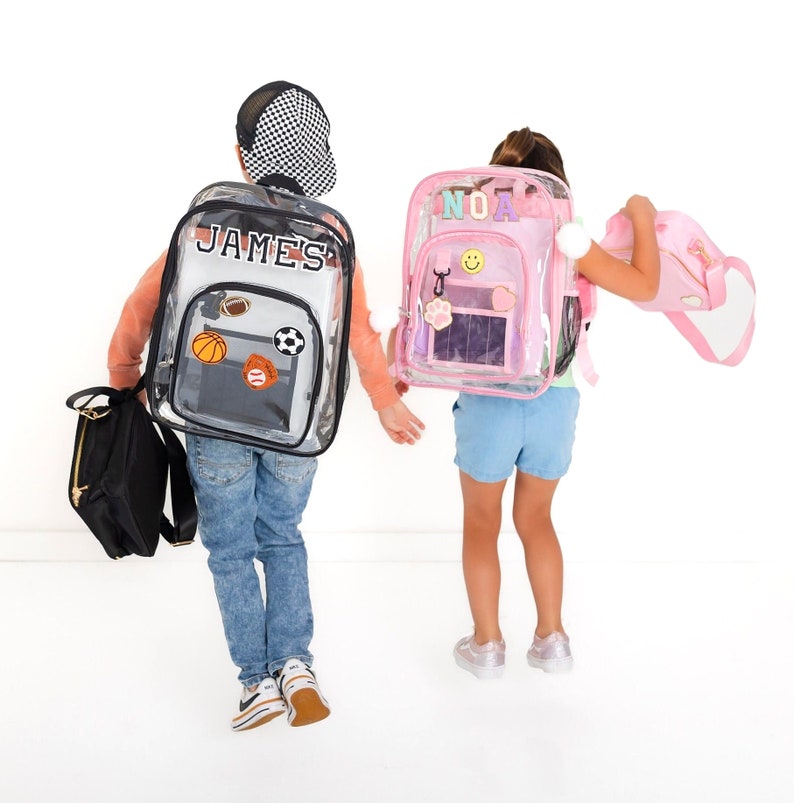 May include: Two clear backpacks with personalized patches. The left backpack features the name JAMES and sports-themed patches. The right pink-trimmed backpack features the name NOA, a smiley face, and a paw print patch, with a matching pink bag.
