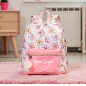 May include: Pink backpack with a floral pattern and the name "Noa Jayne" printed on the front pocket. The backpack features a pink pom-pom zipper pull and a pink handle. The floral pattern includes flowers in pink, yellow, and orange.